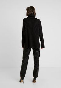 Pull à col roulé noir côtelé, coupe oversize, épaules tombantes, associé à un pantalon cargo noir brillant taille haute et des talons noirs.
