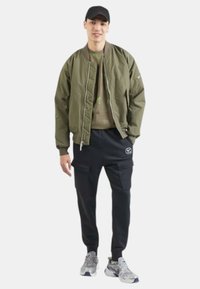 Chaqueta bomber verde oliva sobre una sudadera gráfica beige, combinada con joggers cargo negros y zapatillas grises, con acentos plateados.
