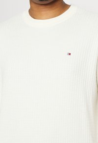 Camicia bianca a maniche lunghe con un motivo a nido d'ape, colletto rotondo e un piccolo logo colorato sul petto.