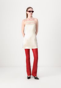 Saténové krémové minidress s priehľadnými vložkami, doplnené červenými slim nohavicami a čiernymi stiletto podpätkami, štýlované s úzkymi červenými slnečnými okuliarmi.