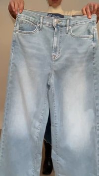 Oseba drži svetlomodre kavbojke z visokim pasom, sprednjimi žepi in gumbno zaponko na enobarvnem ozadju.