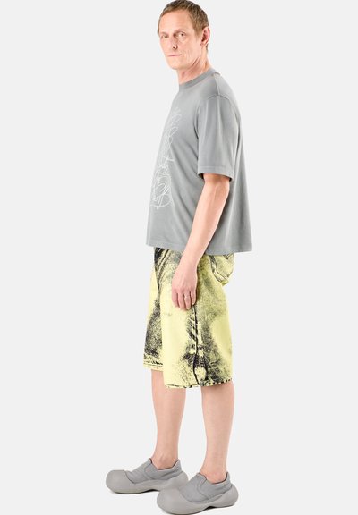 Camiseta gris de gran tamaño con estampado abstracto blanco, combinada con pantalones cortos amarillos con un patrón negro y zapatos gris de estilo slip-on.