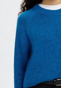 Moderen volnen pulover modre barve z okroglo izrezavo, mehke teksture in sproščenega kroja, kombiniran z belo srajco spodaj.