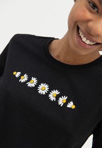 Hymyilevä henkilö, joka on pukeutunut mustaan paitaan, jossa on rinnassa vaakasuora rivi kahdeksasta valkoisesta ja keltaisesta päivänkakkarasta.