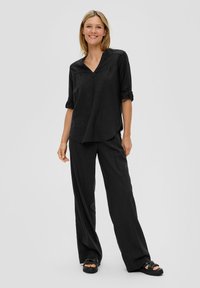 Chemise noire ample avec décolleté en V et manches retroussées. Associée à un pantalon noir à jambes larges. Texture lisse, design décontracté. Sandales noires.