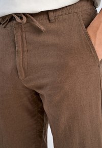 Brązowe spodnie z materiału o miękkiej teksturze, z elastycznym paskiem w talii, bocznymi kieszeniami oraz luźnym krojem.