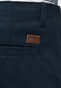 Pantalon bleu marine avec une poche passepoilée à l'arrière et un patch en cuir Timberland marron au-dessus de la poche.