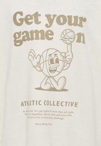 T-shirt bianco in cotone con un personaggio da cartone animato a tema basket, testo beige che dice "Fai decollare il tuo gioco" e "COLLETTIVO ATLETICO" sotto.