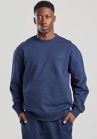 Navy blue sweatshirt in soft material with crew neck, ribbed cuffs, and waistband; features a small, textured logo on the chest.
