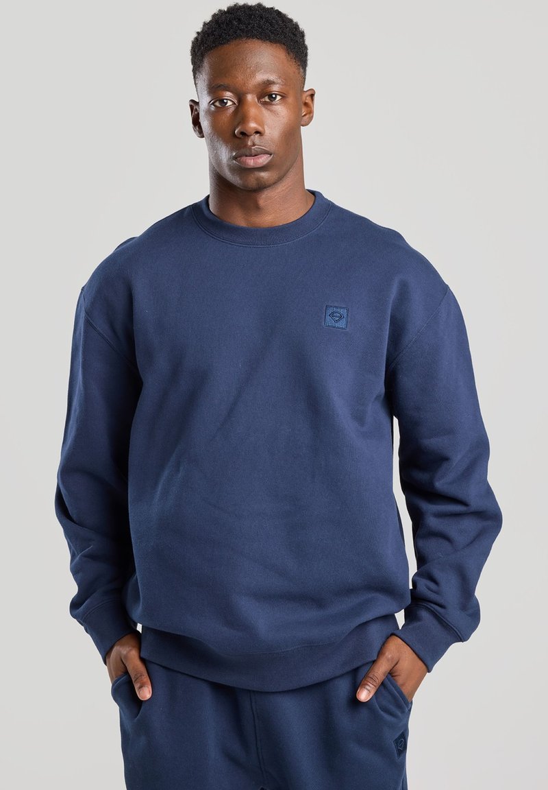 Navy blue sweatshirt in soft material with crew neck, ribbed cuffs, and waistband; features a small, textured logo on the chest.