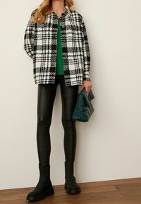 Czarna i biała kratkowana shacket na zielonym topie, w połączeniu z czarnymi skórzanymi legginsami i czarnymi botkami. Trzymając turkusową torebkę kopertową.