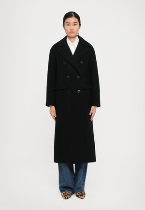 Femme portant un long manteau noir croisé, une chemise blanche, un jean bleu et des chaussures à talons pointus en imprimé léopard, se tenant devant un fond blanc.