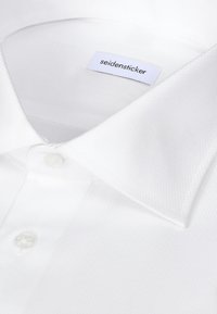 Chemise blanche à col pointu, deux boutons visibles et étiquette de marque "seidensticker" cousue à l'intérieur du col.