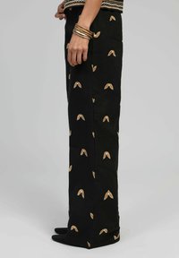 Pantalons noirs à coupe slim, avec des motifs de feuilles beiges brodés, texture lisse et design épuré. Associés à des chaussures noires.