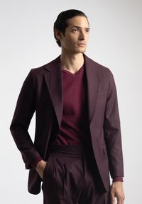Chaqueta de traje color burdeos con solapas de muesca, combinada con un suéter de cuello en V. Ajuste entallado con tejido texturizado visible y bolsillos laterales.