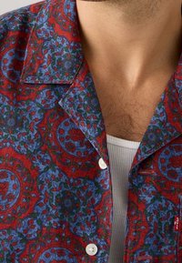 Koszula w czerwono-niebieski wzór z kwiatowym motywem, z kołnierzykiem typu camp i białymi guzikami, wykonana z teksturowanej tkaniny.