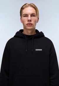 Sudadera negra fabricada en tejido suave, que cuenta con un bolsillo tipo canguro, cordones y un pequeño logotipo blanco en el pecho. Textura lisa.