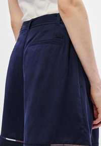 Personne portant un short bleu marine avec des poches arrière et un petit logo de crocodile, et un haut blanc, vu de la taille vers le bas.