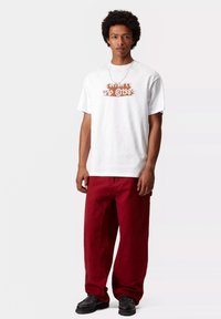 Biała bawełniana koszulka z graficznym wzorem w kolorze brązowym, zestawiona z luźnymi burgundowymi spodniami i czarnymi butami. Model stoi na neutralnym tle.