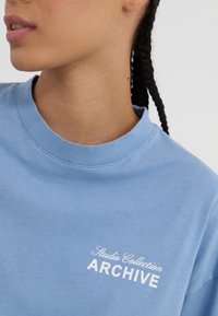Lys blå bomull t-skjorte med rund hals. Har hvit tekst "Studio Collection ARCHIVE" på brystet. Glatt tekstur.