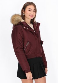 Burgundská péřová bunda s kapucí s kožešinovým lemováním, zapínáním na zip, žebrovanými manžetami a zkráceným designem. Viditelná měkká podšívka.