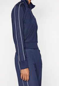 Chaqueta de chándal en navy con cremallera frontal, cuello de canalé y rayas laterales blancas. Confeccionada en una tela suave y elástica con mangas largas.