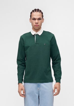 Ung mand med flettet hår iført en mørkegrøn langærmet polo med hvid krave og lyseblå jeans, der står mod en ensfarvet baggrund.