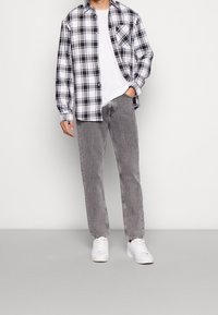Camisa a cuadros en blanco y negro sobre una camiseta blanca, combinada con jeans grises y zapatillas blancas. La camisa tiene un bolsillo en el pecho.