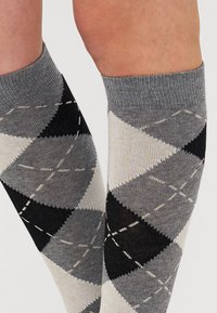 Knee-high kojinės su argyle raštu juodos, pilkos ir kreminės spalvų atspalviuose, su kontrastingais deimantų kontūrais ir šviesiais siūlais.
