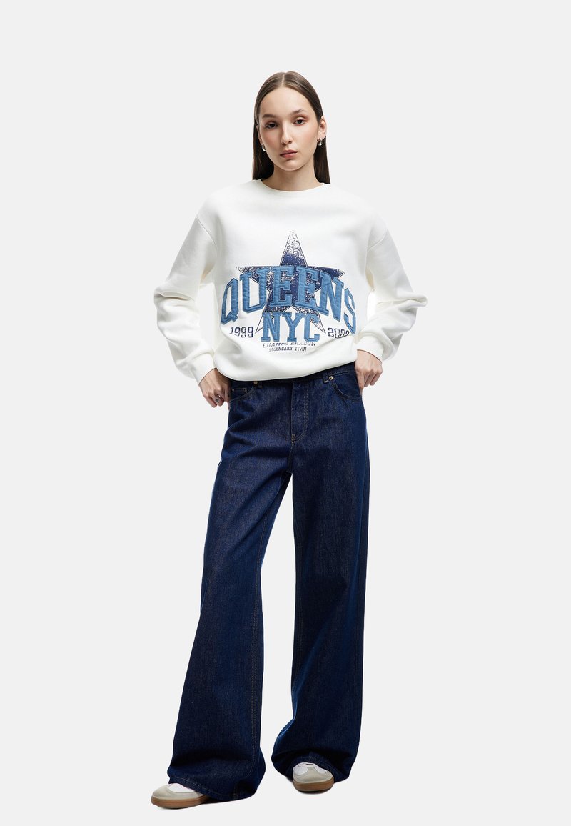Sudadera blanca con estampado gráfico azul y diseño de estrellas, combinada con pantalones de mezclilla oscuros de pierna ancha. La modelo se encuentra en un fondo neutro.