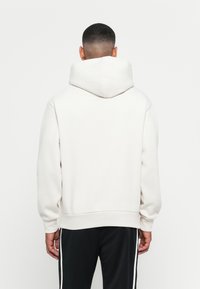 Uomo con capelli corti che indossa una felpa bianca con cappuccio e pantaloni neri con strisce bianche, in piedi con la schiena rivolta verso la fotocamera.