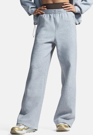 Persoon die lichtgrijze wijd uitlopende joggingbroek en witte sneakers met gouden accenten draagt, handen rustend op de heupen tegen een effen achtergrond.
