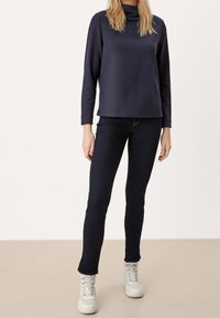 Pullover navy a maniche lunghe con colletto alto, abbinato a jeans skinny in denim scuro e sneakers high-top bianche. Tessuto liscio, design a vestibilità rilassata.