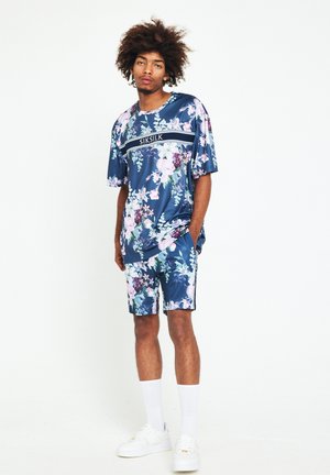 Conjunto deportivo floral en azul marino que incluye una camiseta de manga corta y pantalones cortos, fabricado en un tejido brillante con un acento de logo negro y franjas laterales blancas.