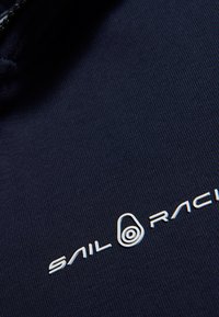 Námorně modrá látka s vertikální žebrovanou strukturou, s bílým logem "SAIL RACING" a kruhovým akcentním designem poblíž loga.