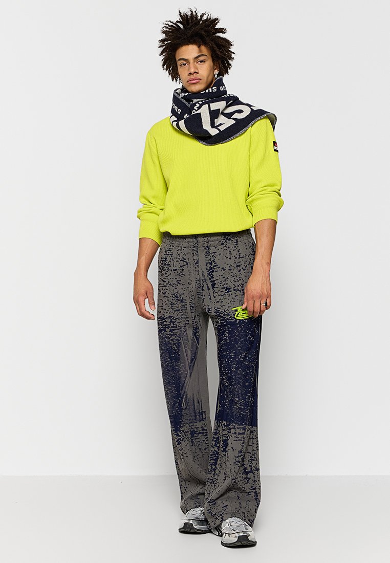 Maglione a coste verde neon, pantaloni larghi a righe navy e grigi, e una grande sciarpa navy con lettere bianche, abbinati a sneakers grigie.