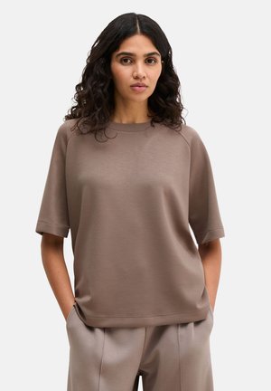Kobieta o falowanych ciemnych włosach, ubrana w luźny taupe krótki rękaw i dopasowane spodnie, z rękami w kieszeniach na tle gładkiego tła.