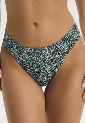 Frau trägt Bikinihose mit hohem Beinausschnitt und schwarz-weißem abstraktem Muster, zeigt Bauch und Oberschenkel vor einfachem Hintergrund.