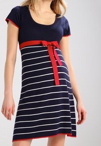 Kvinna iklädd en marinblå och vit randig klänning med korta ärmar, röd dekor och en röd rosett knuten vid midjan.