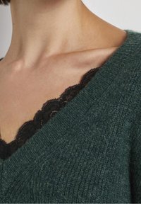 Pull en maille vert foncé avec un décolleté en V, mettant en valeur un tissu texturé. Une dentelle noire de un sous-vêtement est visible.