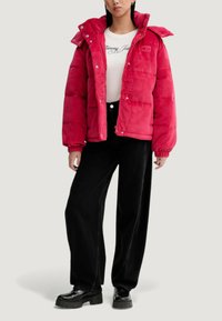Czerwona pikowana kurtka z wysokim kołnierzem, zatrzaskami i elastycznymi mankietami. Noszona na białej koszuli i zestawiona z czarnymi szerokimi spodniami i masywnymi trampkami.