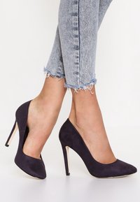 Chaussures à talons aiguilles en daim bleu marine avec un bout pointu et un talon de 10 cm, associées à un jean en denim bleu clair effiloché.