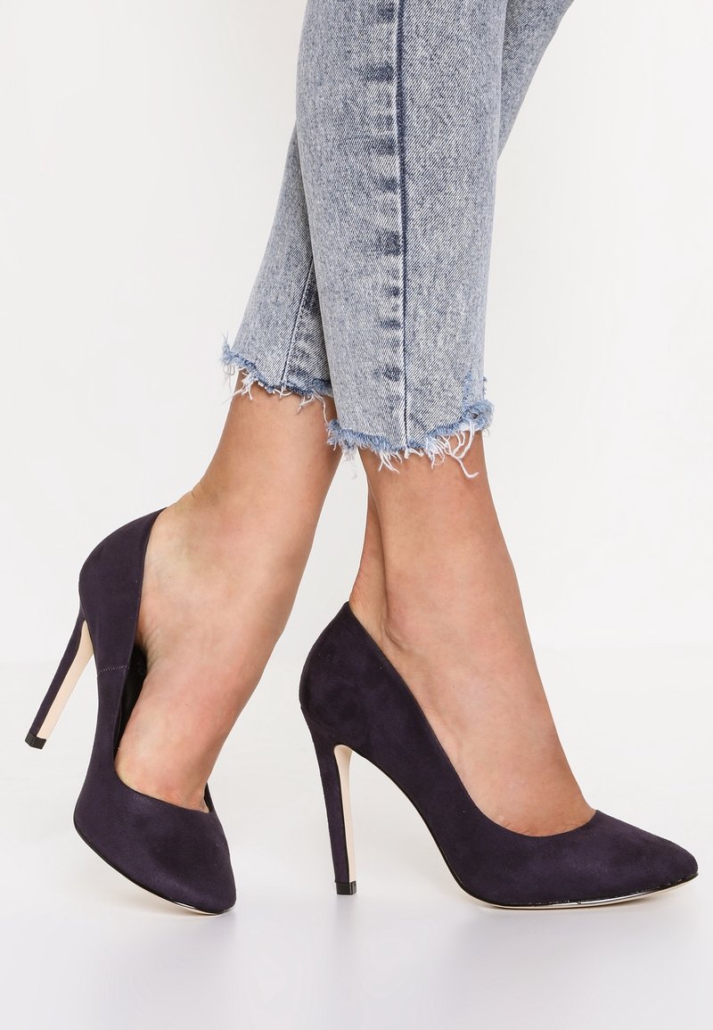 Chaussures à talons aiguilles en daim bleu marine avec un bout pointu et un talon de 10 cm, associées à un jean en denim bleu clair effiloché.