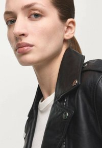 Zwarte leren motorjack met zilverkleurige hardware, gedragen over een wit shirt. Model heeft steil haar en een natuurlijke make-up.