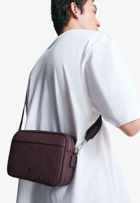 Burgundska crossbody torba s teksturiranim tkanim dizajnom, glatkim naglascima, gornjim zatvaračem i podesivim crnim remenom. Čvrst i kompaktan oblik.