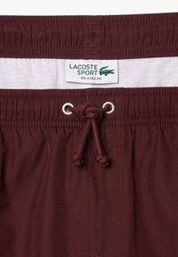Kastanjefärgade shorts med elastisk midja, vit innerfoder, dragsnöre och Lacoste Sport-märke med en grön krokodil-logga.