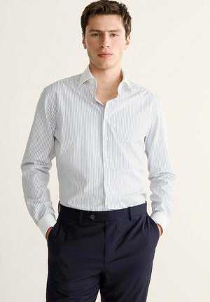 Joven con camisa de rayas blancas y azul claro de manga larga metida en pantalones azul marino oscuro con las manos en los bolsillos.