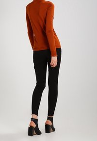 Rostfarbener Langarmtop mit taillierter Passform, kombiniert mit schwarzen Skinny-Jeans. Das Outfit umfasst blockabsatz offene Schuhe.