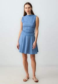 Blauwe, mouwloze jurk met een aangesloten bovenstuk en een uitlopende rok, gekenmerkt door witte stippen. Soepele, lichte stof met een geplooide detail bij de taille.