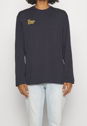 Person wearing a black long-sleeve shirt with yellow text "LIFE AFFIRMING ACTIVISM" on the chest, paired with light blue jeans.